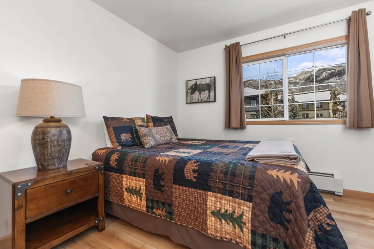 A warm, inviting bedroom scene featuring a bed with a patterned quilt, a window revealing mountain views, a lamp on the nightstand, and framed artwork on the wall. The room is bathed in natural light and designed for relaxing, with elements that evoke comfort and tranquility.