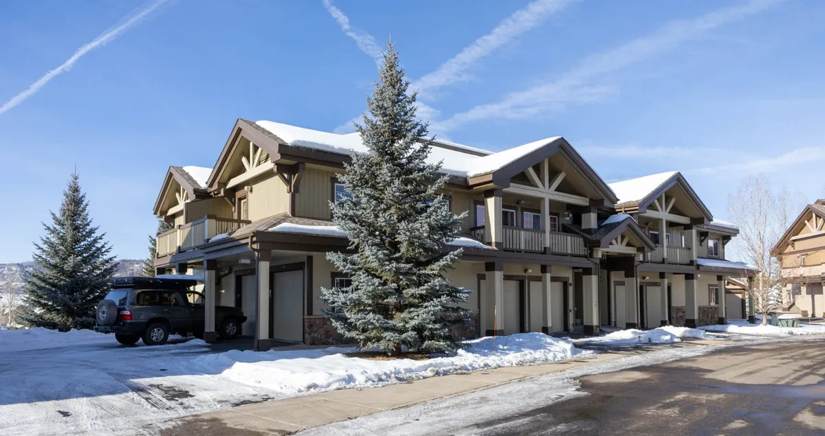 A cozy exterior scene featuring snow-covered roofs and tall evergreen trees surrounding the property, with a well-maintained parking area ideal for storing ski equipment. The inviting look of the building underscores ease of access and a welcoming atmosphere for winter travelers.