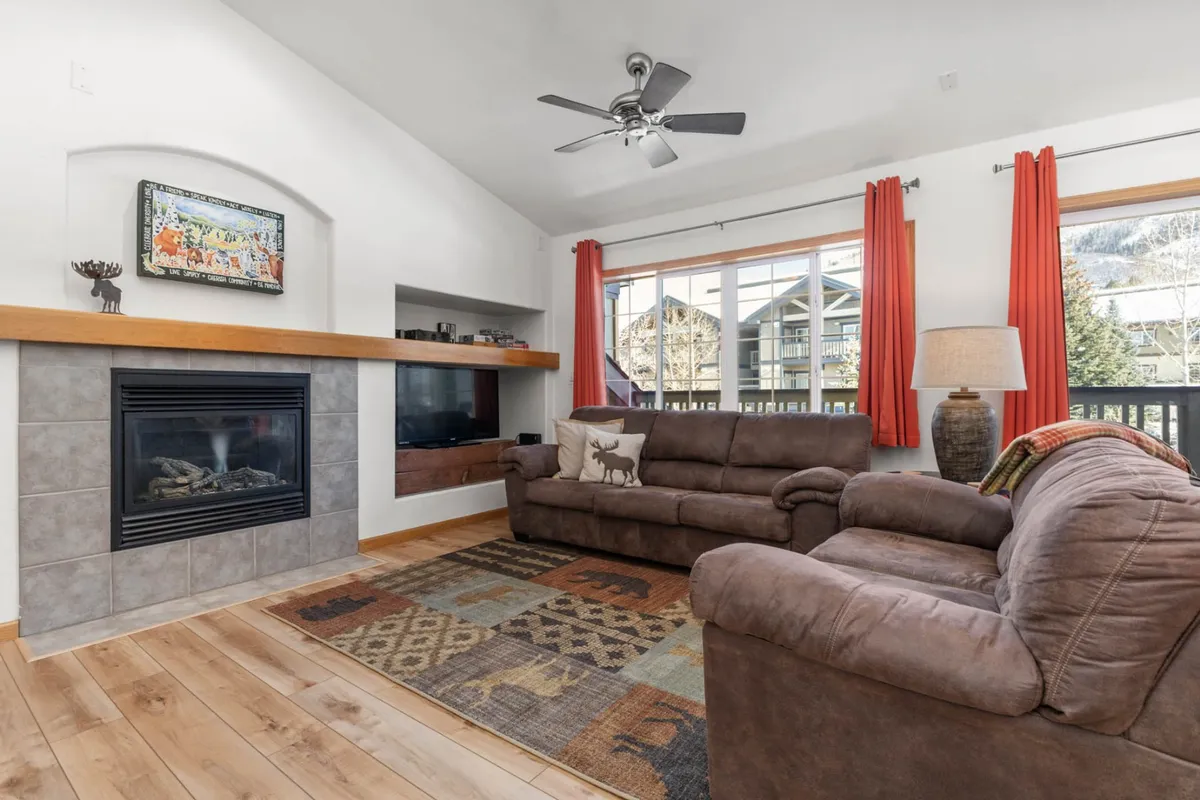 An inviting living space with a fireplace, sofa, large windows, a rug, and decorative art. The mood is welcoming, perfect for relaxing after returning from outdoor pursuits. The windows hint at a connection to the mountains and the outdoors.
