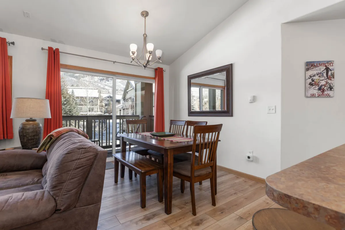 This image features a dining table set near a window, with views to the outside, as well as a cozy sofa, chandelier, and lamp in the background. The scene suggests a space where guests can relax, enjoy meals, or gather after a day of outdoor adventure.