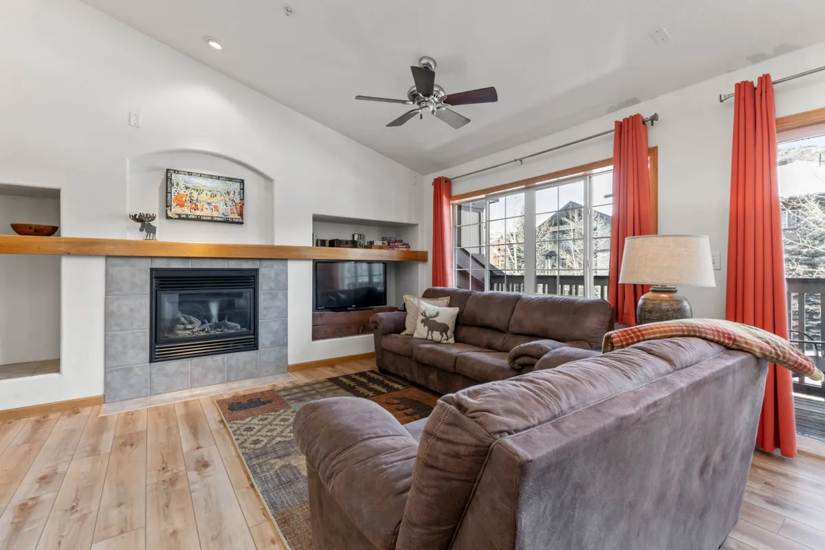 The image showcases a cozy condo living area featuring a crackling fireplace, plush sofa, and expansive windows draped in soft curtains, filling the room with natural light. The inviting setup is perfect for relaxing after a day on the slopes, enjoying quiet mornings with coffee, or gathering with loved ones for memorable moments.