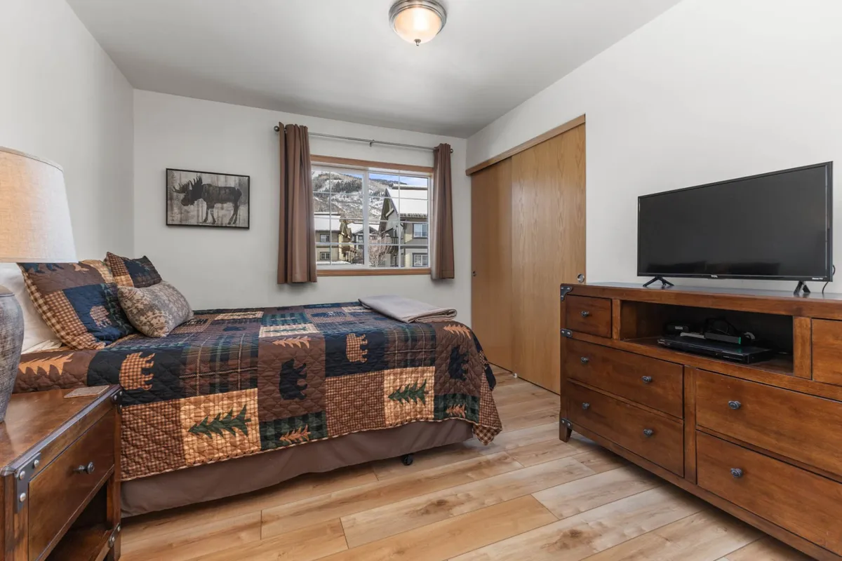 A comfortable bedroom setup with a patterned quilt on the bed, a TV on the dresser, and a window offering a glimpse outside. The space is styled for unwinding, featuring both entertainment and relaxation options.