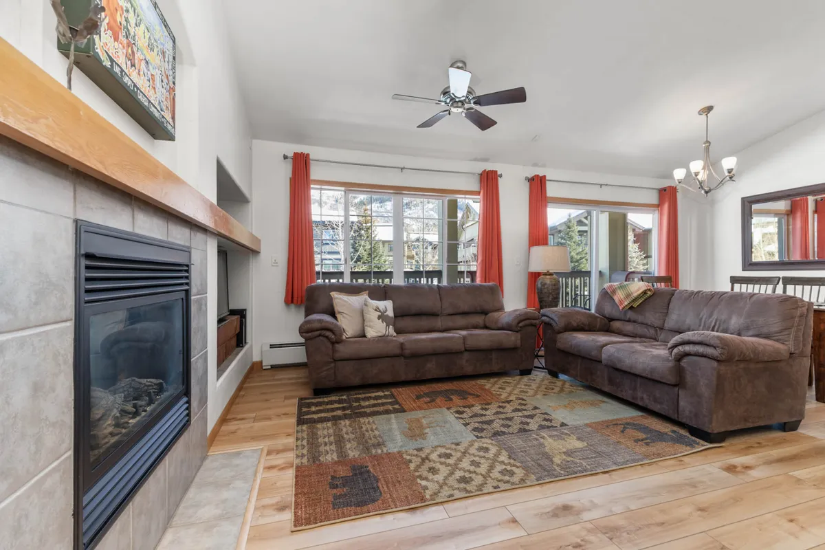 A spacious living room with comfy sofas arranged around a glowing fireplace. Large windows let in abundant natural light, providing a panoramic view of the snowy slopes outside. A fuzzy rug, contemporary decor, and an HDTV complete this inviting gathering area.