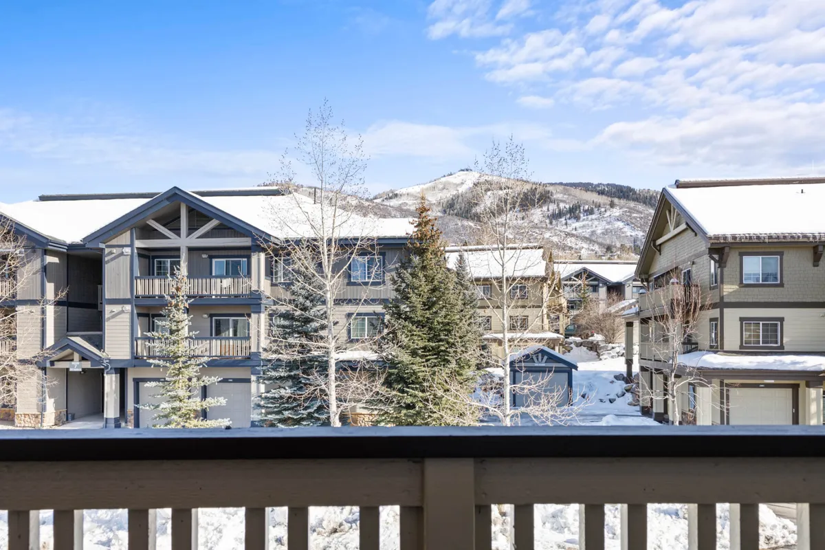 A tranquil winter scene showing snow-covered buildings nestled among pine trees, with picturesque mountains forming a dramatic backdrop. The setting suggests proximity to ski slopes, with visible tracks and hints of winter sports activities.