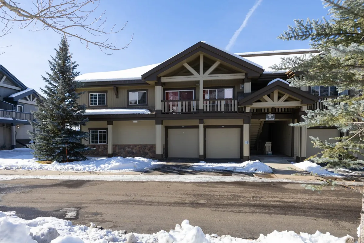 An exterior view of the building showcasing its architectural features, snow-covered grounds, evergreen trees, and multiple balconies. The image captures the inviting and serene atmosphere of the mountain village, with visible access paths that suggest walkability and communal outdoor spaces.